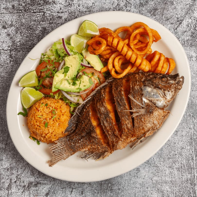 A whole fried fish with Mexican rice, curly fries, and a fresh salad with avocado and lime.