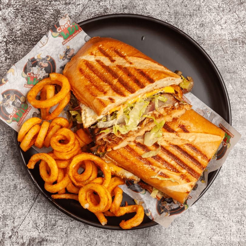 Grilled Mexican torta sandwich with meat, cheese, and lettuce, served with curly fries on a black plate.