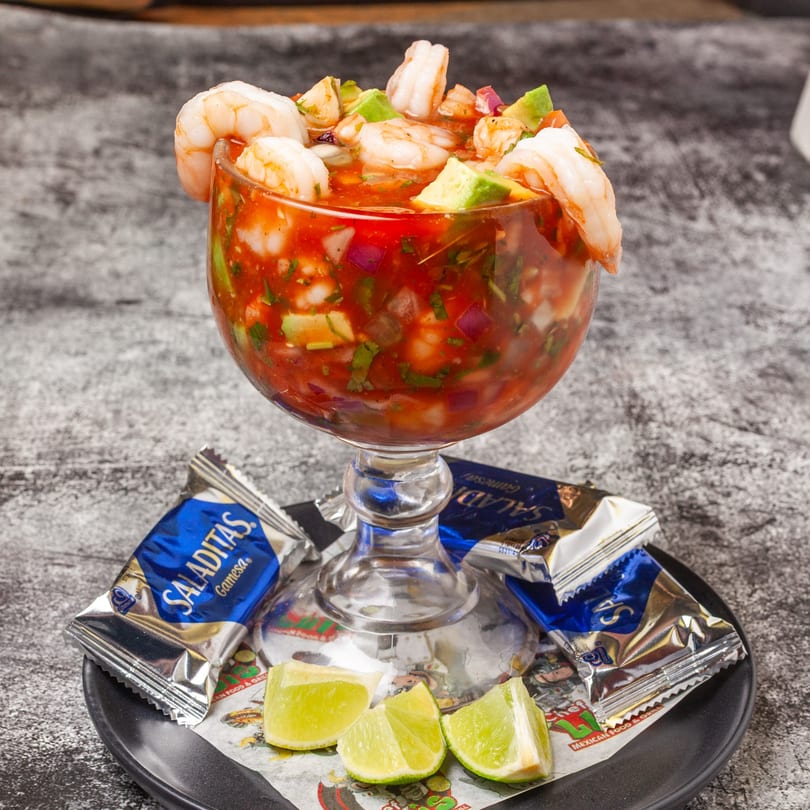 Shrimp cocktail with avocado, red onion, and cilantro in a goblet, served with lime wedges and crackers.